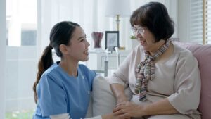 Caregiver assisting an older resident in a bright assisted living common area while another staff member observes.