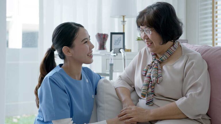Caregiver assisting an older resident in a bright assisted living common area while another staff member observes.